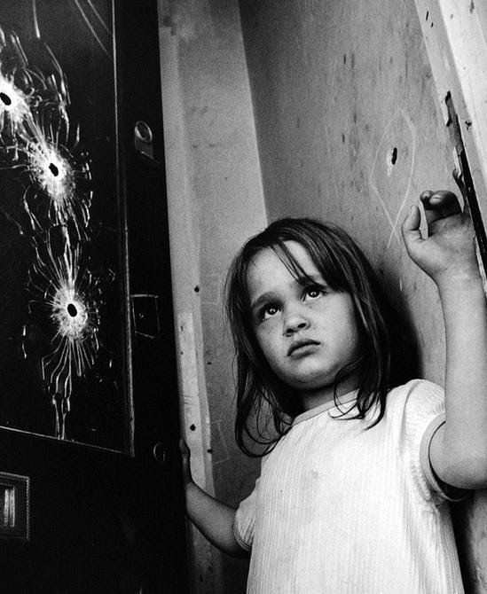 A little girl stands at a front door with gun shots through the window.