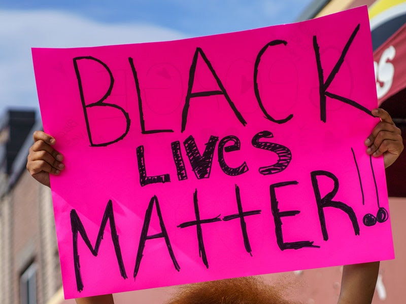 Image of a Black protester holding a pink sign reading in handwritten black lettering %22BLACK LIVES MATTER!!%22 at a street protest