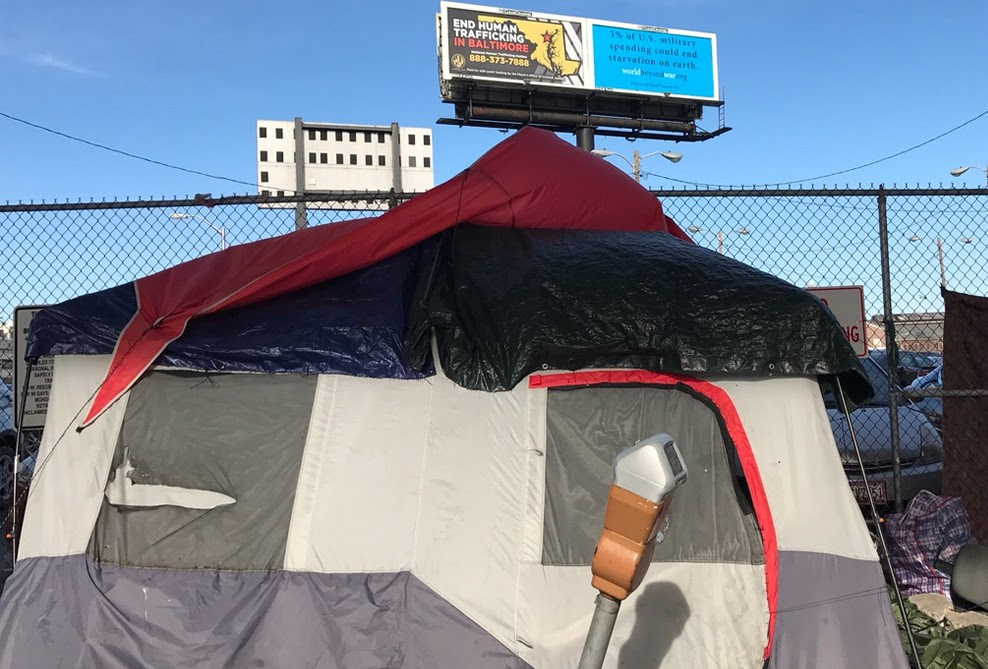 Homeless encampment in the foreground of a Baltimore, MD billboard that read, “3% of US military spending could end starvation on earth.” Source World Beyond War.