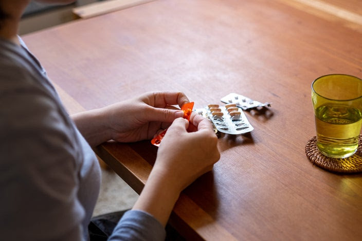 Close-up of a woman's hand taking decongestant pills out of a blister pack with a glass of water on the table with her.