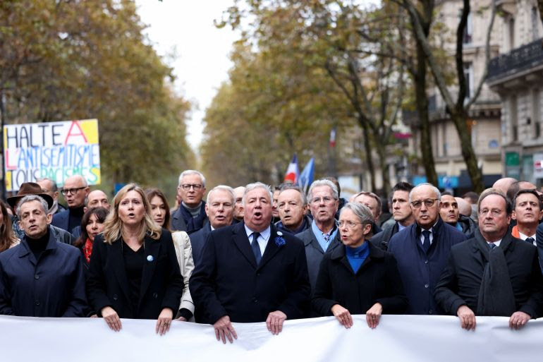 Senior French politicians including President of the National Assembly Yael Braun-Pivet, Prime Minister Elisabeth Borne, Senate President Gerard Larcher and former presidents Francois Hollande and Nicolas Sarkozy at the front of the march. They are walking behind a banner. Someone behind them is holding up a placard reading 'Stop anti-Semitism and all racism'