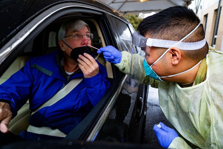 A healthcare professional wearing PPE administers a COVID-19 test to a patient in his car