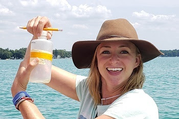 a smiling young woman with shoulder-length light brown hair and a wide-brimmed brown hat holds a small plastic bottle and pencil, water in background