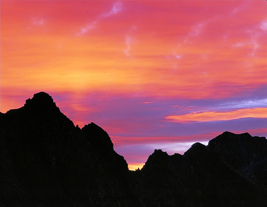 Silhouetted mountain landscape against pink-orange sunset by John Fielder Silhouetted mountain landscape against pink-orange sunset by John Fielder