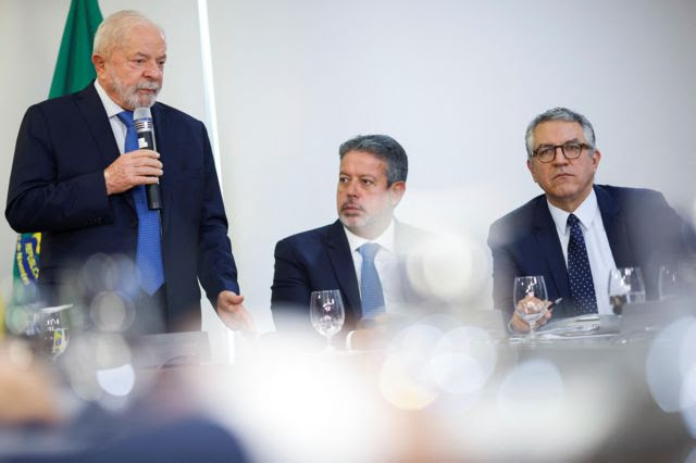 O presidente Lula fala ao microfone ao lado do presidente da Câmara, Arthur Lira, e do ministro das Relações Institucionais, Alexandre Padilha