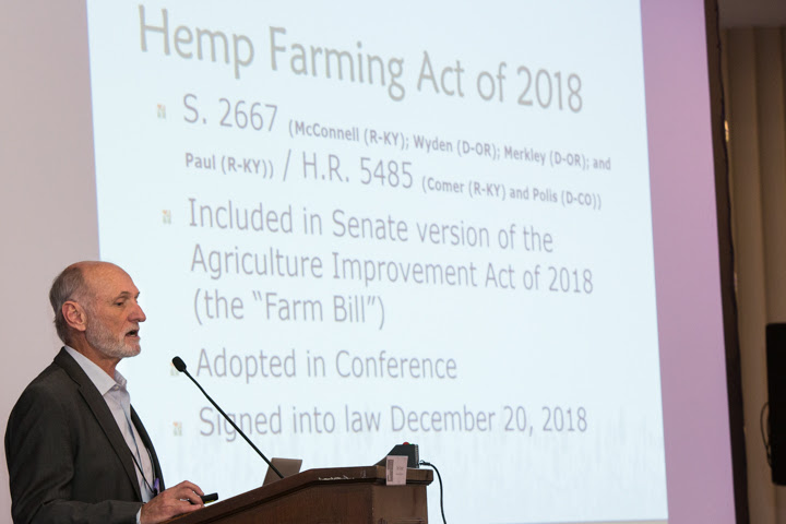 Michael Mcguffin, and older white man with gray hair and a short
beard is speaking, standing at a podium in front of a projection on a
screen. The projection is of a slide presentation and the slide
displayed is about the Hemp Farming Act of 2018.