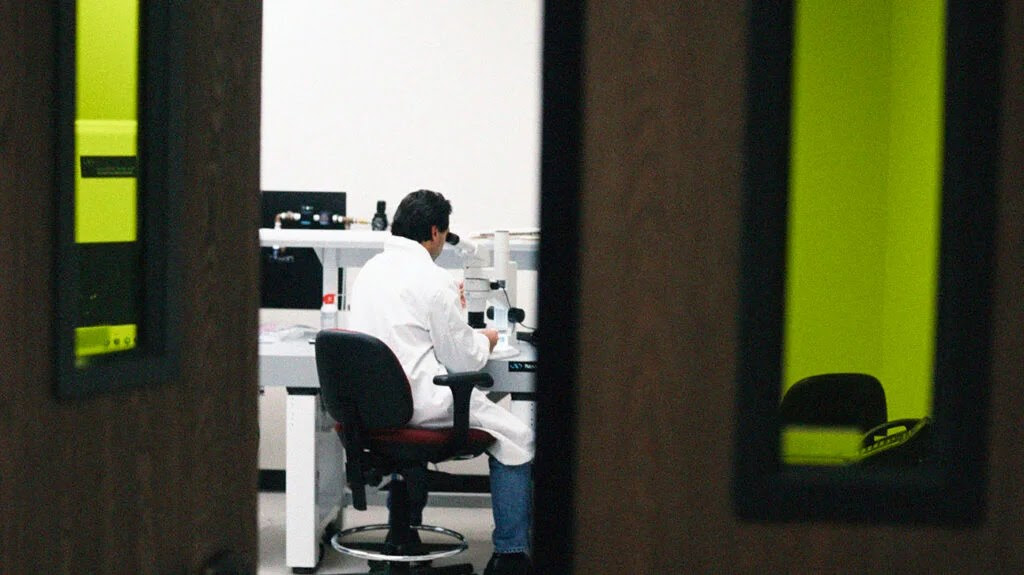 A researcher wearing a lab coat looks into a microscope inside a lab