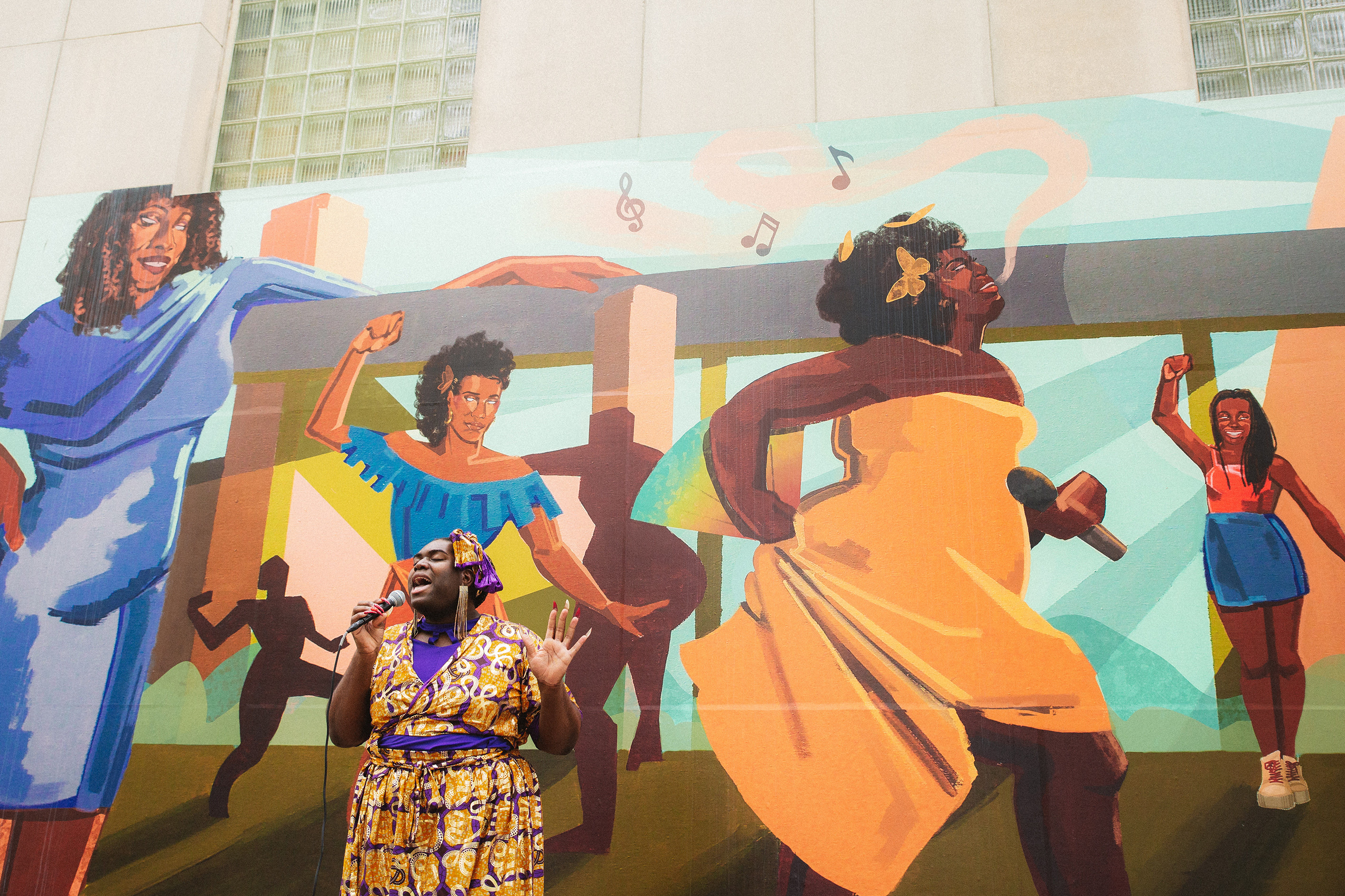 Qween Jean is holding a microphone, wearing a orange and purple dress with a matching headwrap, and long earrings. She is standing in outside of the Queens Museum, in front of Glori Tuitt's mural "Black, Trans & Alive "Qweens Song)"