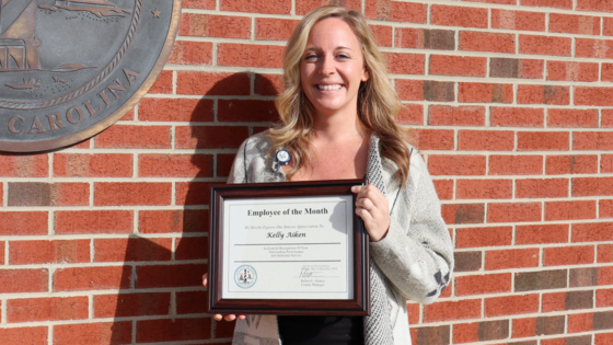 Image of Kelly Aiken holding her certificate standing in front of a brick wall.
