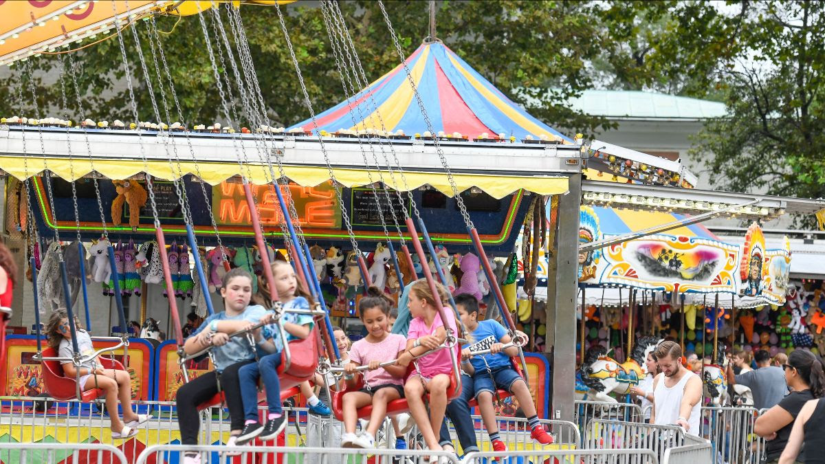 Children ride a carnival ride. In the background crowds of people play carnival games and walk around a fairground.