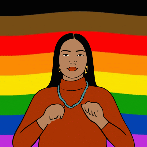 An olive-skinned femme-presenting person with hoop earrings, in front of a  “More Color, More Pride” flag, tracing a heart in the air with their fingers.