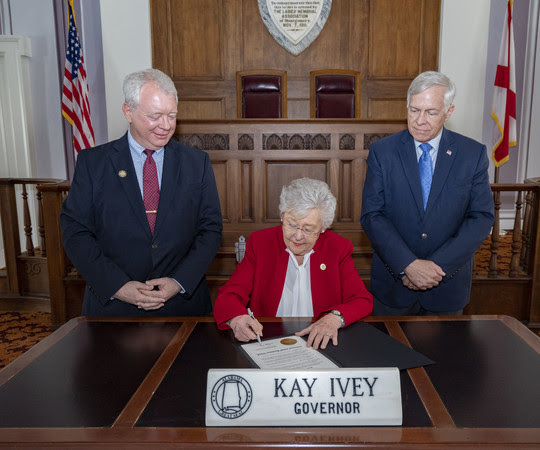 Governor Kay Ivey signs proclamation with district director standing on the right and deputy district director stand on the left watching.