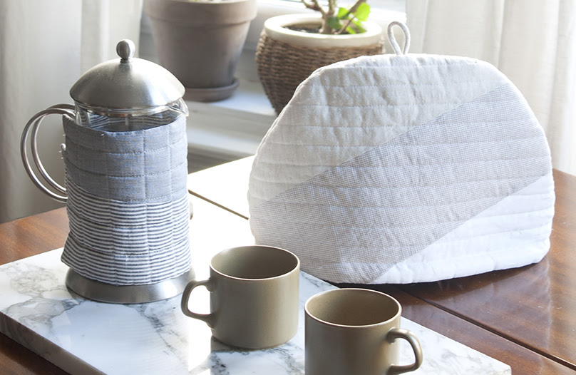 A photograph of a tea and coffee set on a wooden table, by a window. On the foreground there is a french press and two beige mug on a marble plate, the french press is wrapped in a quilt warmer by Woolgathering. Behind, there is a teapot covered by a quilt warmer by Woolgathering. There is plenty of sunlight and plants at the window.