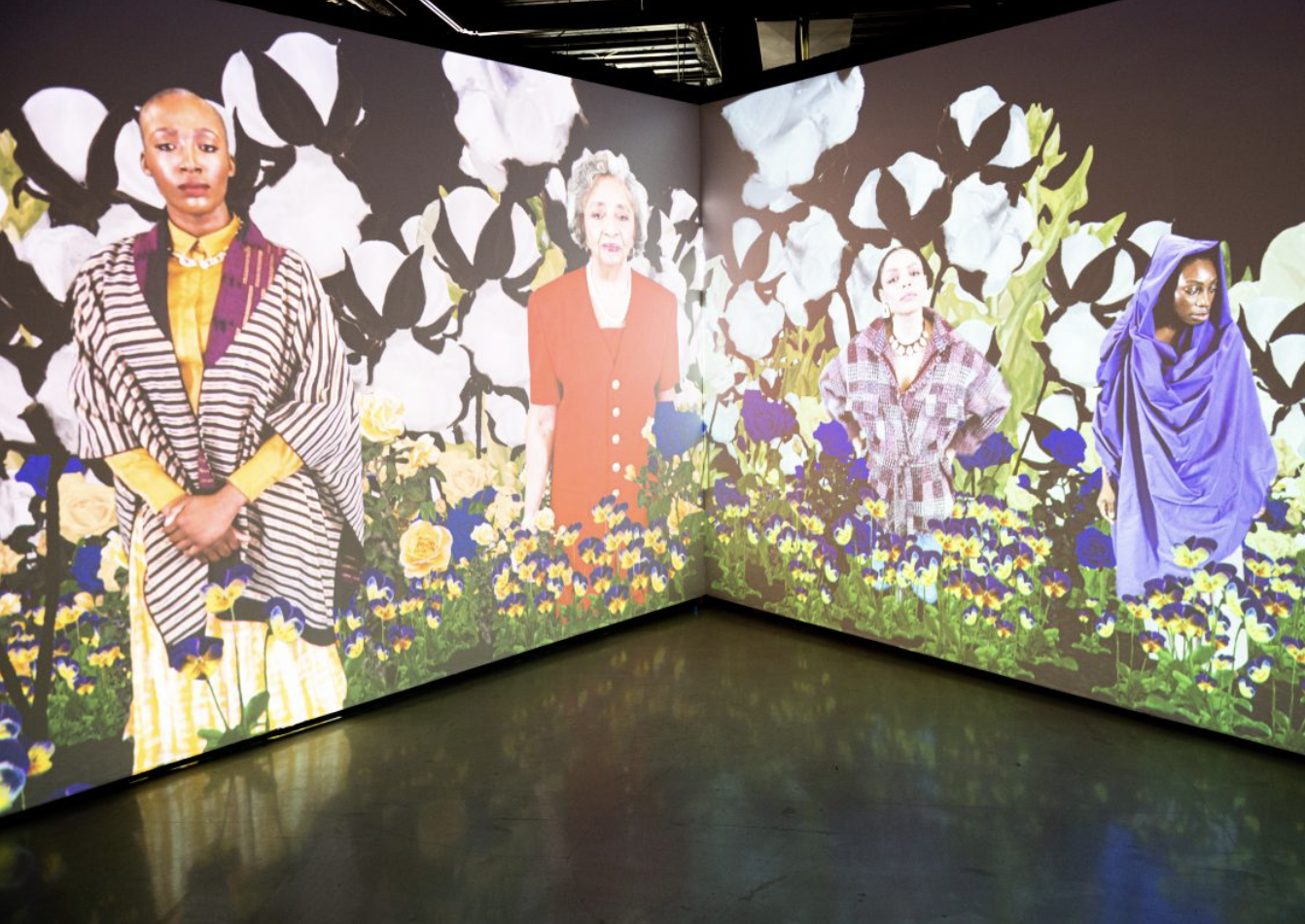 An installation view of Stephanie Dinkins "Secret Garden" (2021) at Stamps Gallery, University of Michigan. The interactive video projection shows four generations of black women standing in a garden where cotton, roses, and okra grow. 