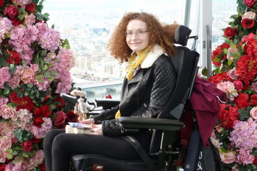 Hannah Deakin with red curly hair in her wheelchair wearing black trousers and a black leather jacket in front of an archway of pink and red flowers
