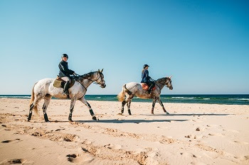 two women in riding gear sit atop two cream-colored horses walking along a sandy beach next to a huge, calm, blue lake