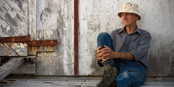 A man wearing a fishing hat is sitting down leaning against against a weathered wooden building.