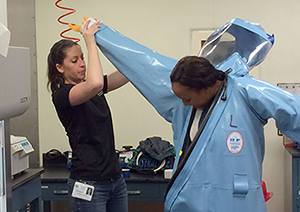 An instructor assists a participant into a BSL-4 protective suit before conducting simulated BSL-4 lab exercises during the 2015 data quality course