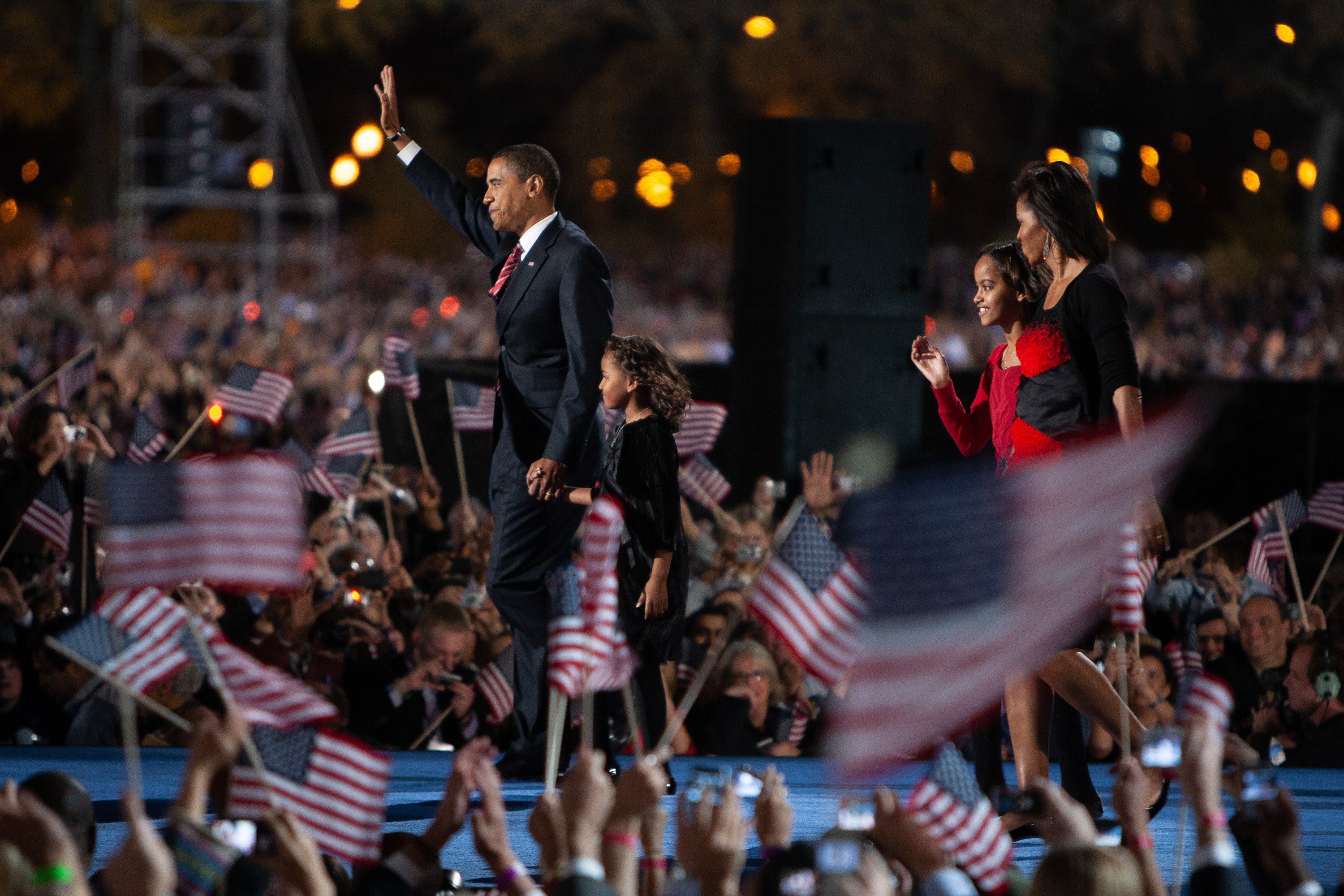 President Obama raises his hand as he walks across a blue stage. He is holding hands with Sasha Obama. Behind him is Michelle and Malia Obama. They are surrounded by a crowd waving American flags below. 