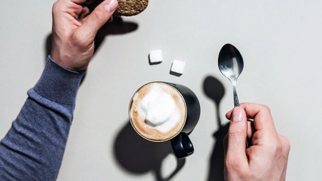 photo showing hands holding a cookie and teaspoon, with a foamy drink in a black mug at the center of the photo and two sugar cubes next to it