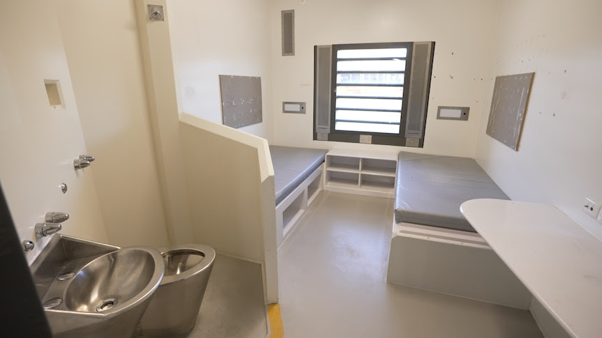 A small room with two bare single beds on either side, with a stainless steel basin and toilet in the foreground.