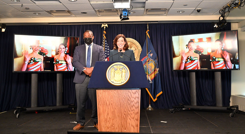 Governor Hochul and Lieutenant Governor Benjamin stand at podium with two video screens displaying the Rockettes