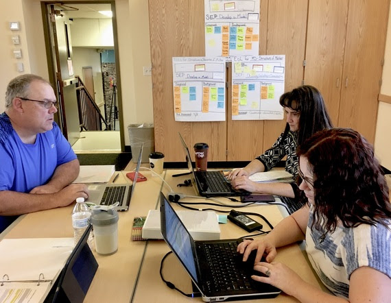 Three educators sit around a table working intensely on their laptops. The wall behind them is full of posters covered with sticky notes from the workshop.