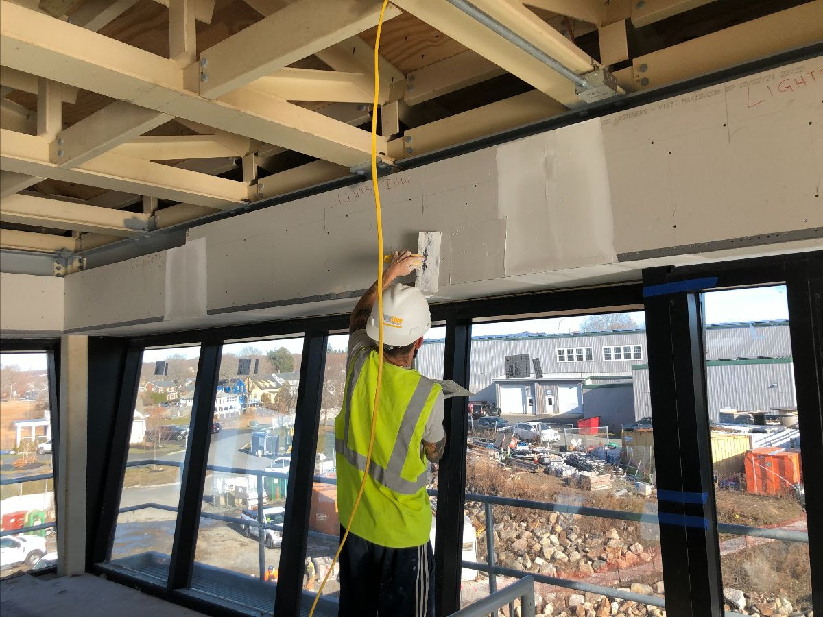 A worker in a yellow vest and hardhat smooths plaster over the joins in a span of drywall above windows in the control tower. The windows overlook the construction site below. The wooden beams below the ceiling of the control tower can be seen above the worker's head. 
