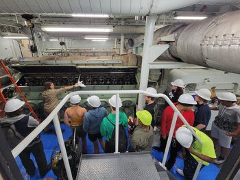 Several young adults watch on as a ferry engine room crewmember shows an engine and talks to them