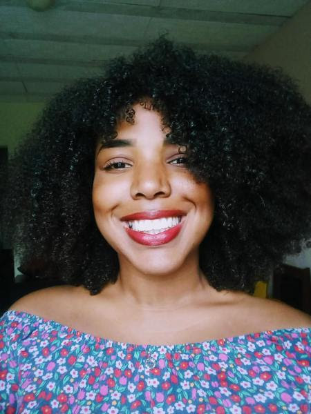 Ana Paula, atual estudante do prouni. Ela é uma mulher negra com cabelo cacheado escuro na altura dos ombros. Ana está sorrindo para a câmera. Ela usa batom vermelho e usa ombro a ombro florida
