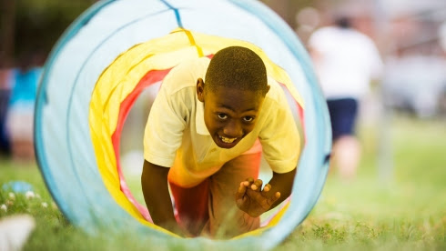 A boy is crawling through a bright fabric tunnel in an outdoor setting.