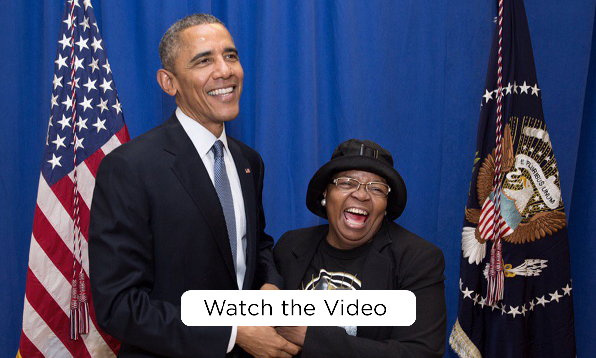 President Obama smiles as holds the hand of Edith Childs, a Black woman with a medium deep skin tone. She is smiling, wearing glasses, and a black bucket hat. In the background is the American flag and the Flag of the President of the United States. At the bottom of the image, a white rectangular button reads, “Watch the video.” 