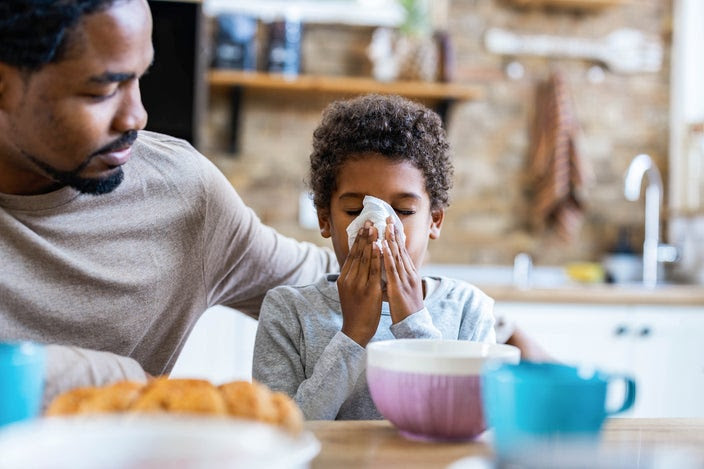 Portrait of a young boy blowing his nose with his father sitting next to him at the kitchen table. The father is comforting the young boy with his arm around his son's shoulders.