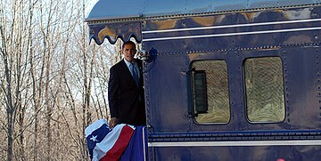 Obama is standing aboard the back platform of a train and looking to the side of the train. There is a red, white and blue banner hanging over the rear railing.