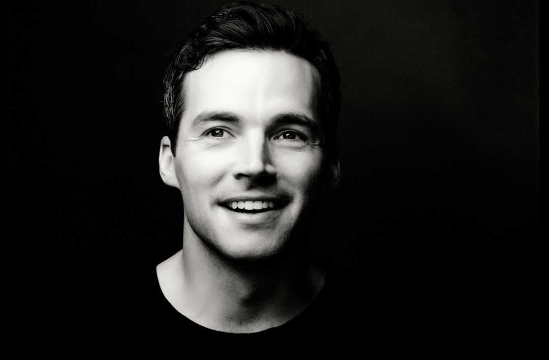 A black and white photo of Ian Harding, a man with short brown hair and a black shirt. He is smiling.