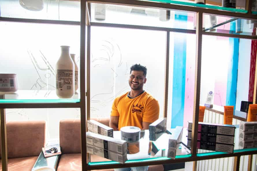 A man in a yellow t-shirt smiles from behind shelves in the salon
