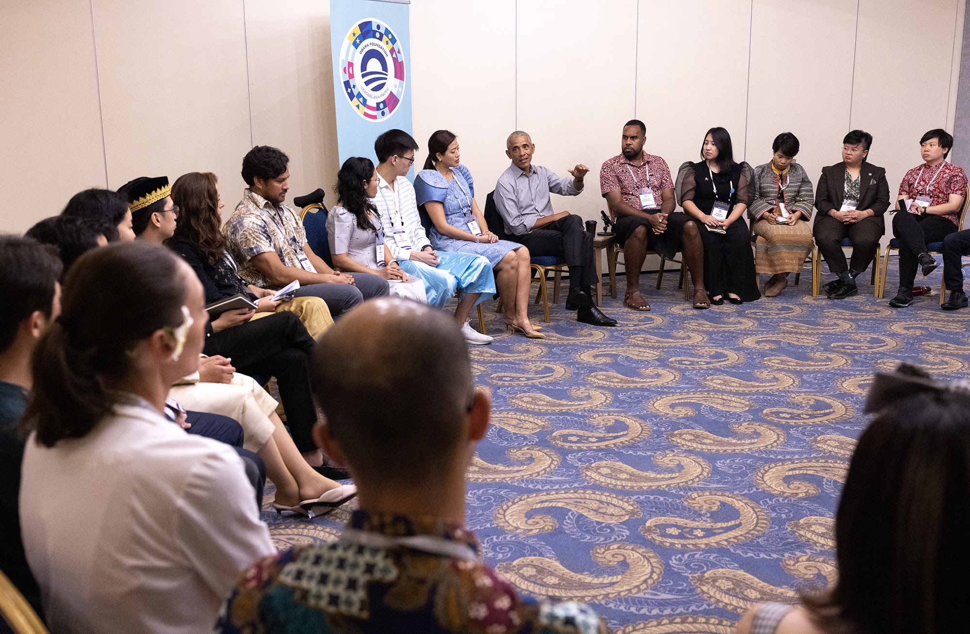 President Obama is sitting in a chair wearing a collared shirt, mid sentence with his arm raised. A group of people with a range of skin tones and hair types are seated around him in a semicircle, listening to him speak. 