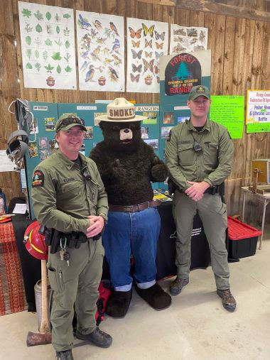 Two Rangers and Smokey Bear stand in front of an informational table at a county fair