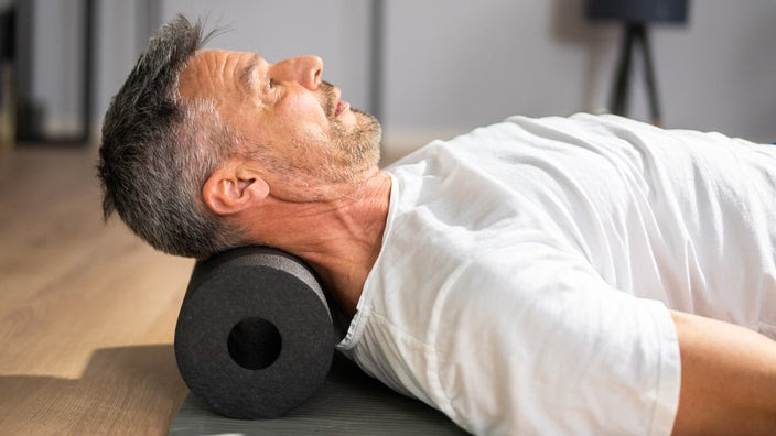 A man lies on his back with a foam roller behind his head. It rests in the arch of his neck.