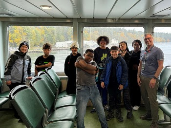 Several people posing for a photo on inside the passenger cabin of a ferry with windows looking out in the back of them