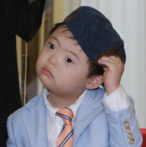 toddler-aged boy wearing a cap and jacket and tie with a bewildered look on his face