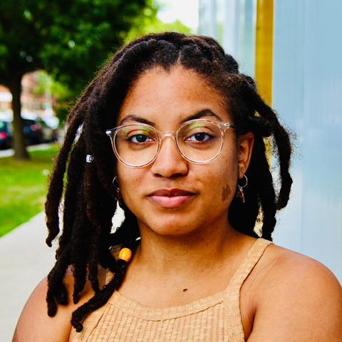 A light-skinned Black femme from the chest up with clear-framed glasses, small gold dangly earrings, shoulder length locs, and brown birthmark on her left cheek.