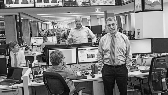 Black-and-white photo of Martin Baron standing in Washington Post newsroom near working reporters, desks, and screens