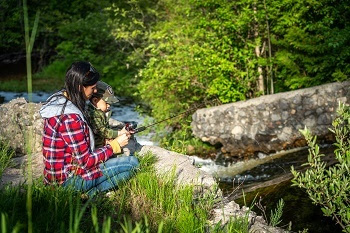 a woman with long dark hair, wearing a red plaid jacket, helps a young boy wearing a baseball cap hold a fishing pole out over a grassy, rocky ridge