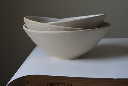 A photograph of three stacked beige ceramics bowls by Hazel Frost. They are displayed against a beige wall and on a white piece of paper and wooden table.