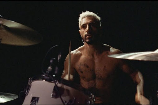Ruben from the film Sound of Metal with bleach blonde hair sat in front of a drum kit topless