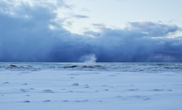 a start, white and gray snow-covered shoreline, with a wall of swirling dark gray clouds and hints of sunlight just behind it