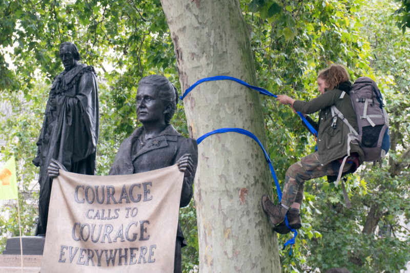 Protestor climbs a tree in Parliament Square in September. Statue with sign "Courage Calls to Courage Everywhere"