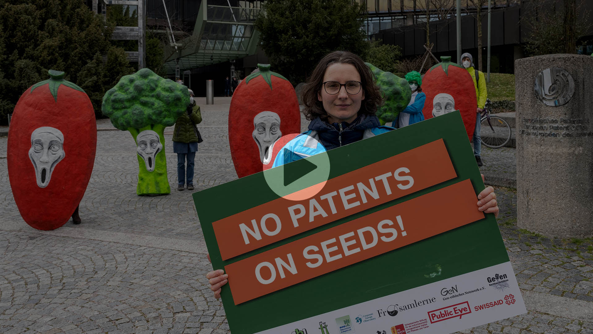 Screenshot of a video, a woman stands in centre with No Patents sign, some people in line behind her dressed as vegetables, play button in centre