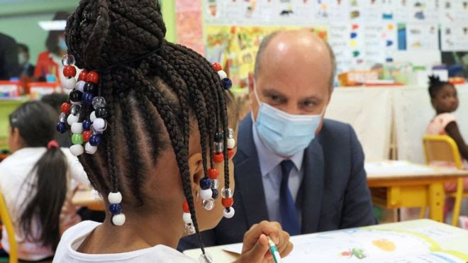 O ministro de educação da França, Jean-Michel Blanquer, de máscara, conversa com uma menina de tranças no cabelo, sentada em sua carteira, na escola